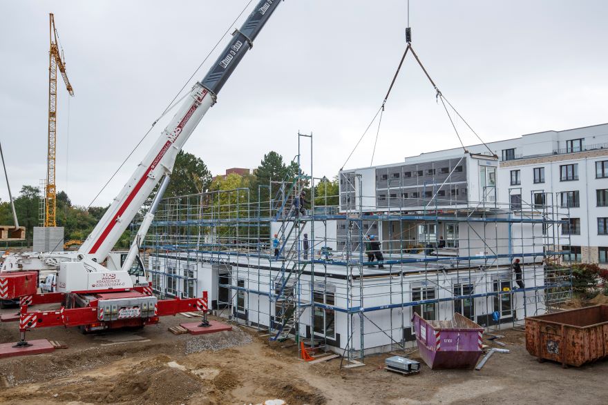 Ein Raumodul schwebt am Hacken des Krans über der Baustelle.